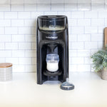 Eine schwarze Babybrezza.de Formula Pro Advanced inkl. zusätzlichem Milchpulvertrichter steht auf einer Küchentheke mit weißer Fliesenrückwand. Unter dem Ausguss befindet sich eine mit Milchpulver gefüllte Flasche, daneben ein Sauger für eine Babyflasche. Eine Pflanze und ein gemusterter Behälter unterstreichen die ruhige Szene im Hintergrund. - product thumbnail