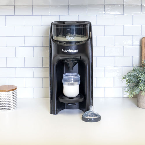 Eine schwarze Babybrezza.de Formula Pro Advanced inkl. zusätzlichem Milchpulvertrichter steht auf einer Küchentheke mit weißer Fliesenrückwand. Unter dem Ausguss befindet sich eine mit Milchpulver gefüllte Flasche, daneben ein Sauger für eine Babyflasche. Eine Pflanze und ein gemusterter Behälter unterstreichen die ruhige Szene im Hintergrund. - product thumbnail