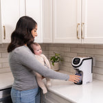 Eine Frau mit einem Baby nutzt den Formula Pro Mini: Babyflaschen unterwegs zubereiten von Babybrezza.de, mit bewährter Mischtechnologie. Sie drückt einen Knopf am kompakten, mobilen Gerät neben weißen Schränken und einer Topfpflanze. - product thumbnail