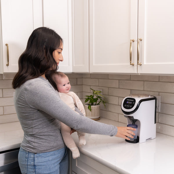 Eine Frau mit einem Baby nutzt den Formula Pro Mini: Babyflaschen unterwegs zubereiten von Babybrezza.de, mit bewährter Mischtechnologie. Sie drückt einen Knopf am kompakten, mobilen Gerät neben weißen Schränken und einer Topfpflanze. - product thumbnail