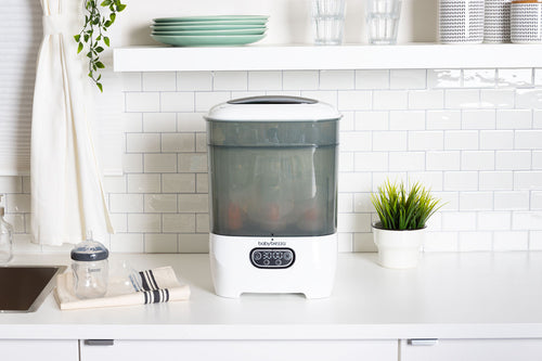 Der Babybrezza.de Sterilizer Dryer Advanced: Automatischer Babyflaschen-Sterilisator steht auf einer weißen Küchentheke neben einer Flasche, einem Handtuch, einer Pflanze und Glaswaren – ideal für die praktische Sterilisation von Babyflaschen. - product thumbnail
