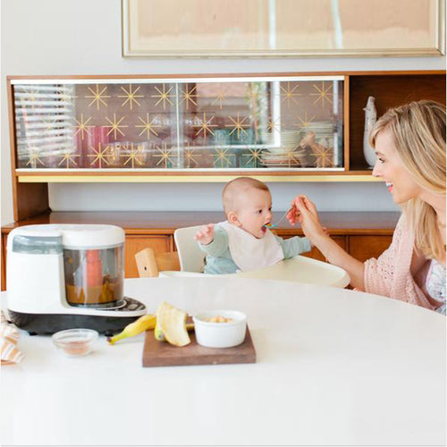 Eine Frau lächelt, während sie ein Baby im Hochstuhl an einem weißen Tisch füttert. Daneben steht ein Baby Brezza Food Maker Deluxe von Baby Brezza.de neben einer Schüssel und Bananen bereit. Ein Retro-Schrank mit Sternenmuster verleiht dieser spülmaschinenfesten Küchenszene Charme. - product thumbnail